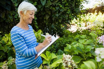 Smiling senior woman writing on clipboard while standing amidst