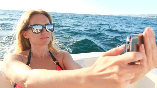 An attractive woman on a yacht on a summer day at sea makes herself on her smartphone. a thin busty blonde with long hair, wearing sunglasses . 4, copy space, close-up. Slow motion