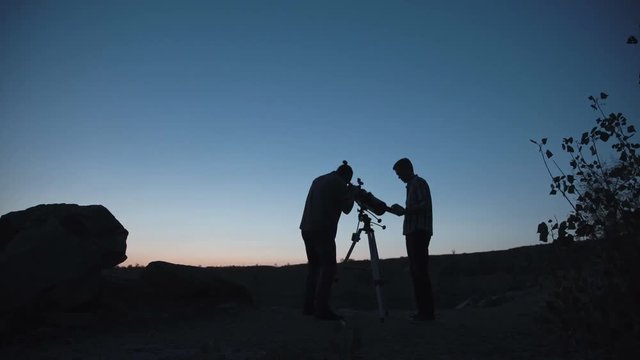 Wide view of silhouettes of people looking through telescope on shore of lake in dark.