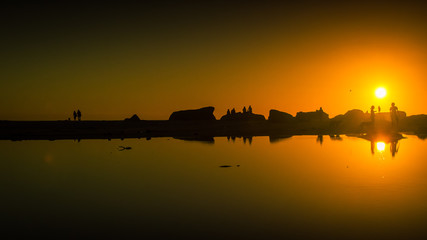 Families walk and play on the beach at sunset, reflected in the still water of the lagoon.