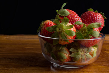 Strawberries in glass bowl on wooden tray against black background.
