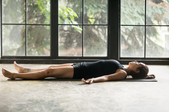 Young attractive woman practicing yoga at home, lying in Savasana exercise, Corpse pose, working out, wearing sportswear, black shorts and top, indoor full length, studio background