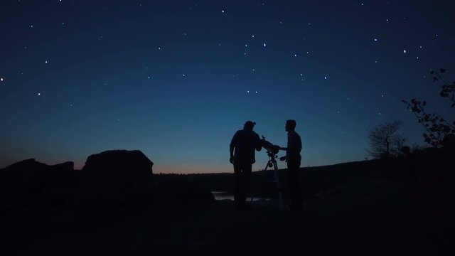 Black silhouettes of people looking through telescope on stars in dark twilight on shore.