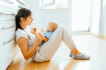 Cozy bright picture of happy laughing mother in pajamas sitting with her baby boy on the floor at the kitchen at home. Parenthood, happy family and sweet home concept.