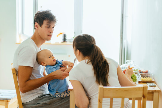 Real people scene: young happy family having breakfast together. Infant baby sitting on father hands and feeding his mother with fork. Routine, way of life and family relations concept.