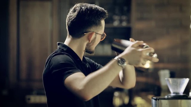 Trendy bearded bartender dressed in black uniform is shaking bottle preparing beverages in night club. Stylish man is working in the pub mixing ingredients in shaker for making tasty fresh cocktails.