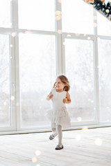 Little happy girl in a festive white dress, looks out the panoramic loft window, dances, decorates a Christmas tree in a pot with a garland of lights bokeh.  Winter rest, waiting miracle of New Year