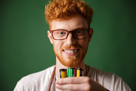 Close-up portrait of readhead bearded photographer in glasses, holding camera rolls, looking at camera