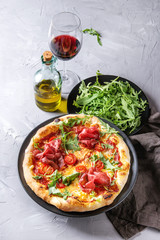 Whole homemade pizza with cheese and bresaola, served on black plate with fresh arugula, olive oil, glass of red wine and textile napkin over gray concrete texture background.