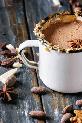 Vintage mug of hot chocolate, decor with nuts, caramel, spices. Ingredients above. Chopped dark and white chocolate, cocoa beans, anise over old wooden table. Dark rustic style. Close up