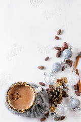 Vintage mug in wool scarf of hot chocolate, decor with nuts, caramel, spices. Ingredients and Christmas toys above over white texture background. Top view with space.