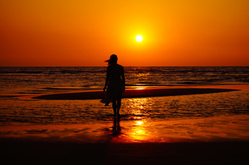Fototapeta premium Woman stand on the beach and look to the sunset. Female in a dress on the beach. Woman silhouette.