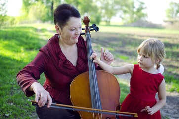 Grandmother and granddaughter playing outdoors on cello. Adult woman teaching girl to play violocello