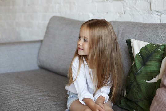 Little girl with charming smile and long straight hair sitting on grey couch and looking sideways at something with interest. Cute female child spending time at home, waiting for brother form school