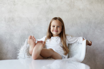 Cheerful pretty little girl with charming smile having relaxed and carefree look, spending weekend morning at home, sitting on white armchair, resting bare feet on table, ready to watch cartoons