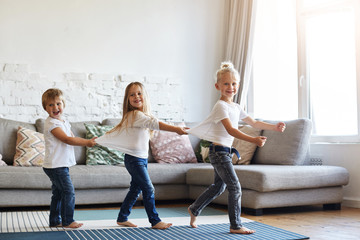 Candid shot of three cute children wearing similar white t-shirts and blue jeans walking barefooted on floor at home while dancing or doing conga line indoors on birthday party, having happy faces