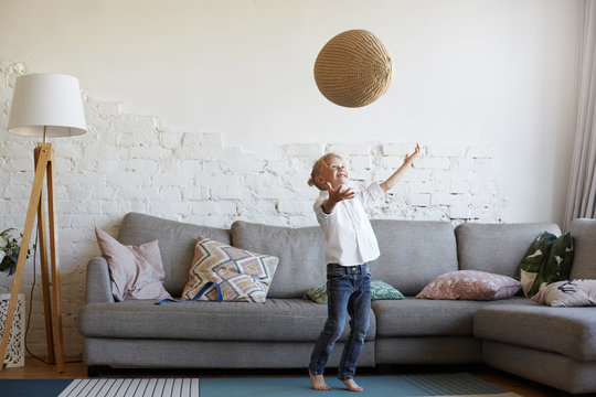 Happy active blonde little boy in white shirt and jeans standing at grey couch in modern home interior, playing with woven ball, throwing it in the air and then catching, having joyful cheerful smile