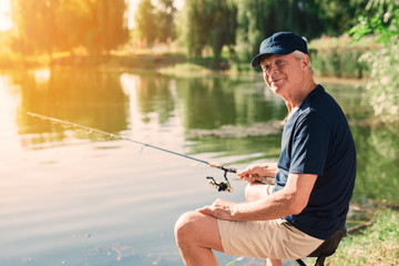 The old man sits on the bank of the river with a spinning in his hands and catches fish. He is looking at the camera