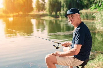 Gordijnen The old man sits on the bank of the river with a spinning in his hands and catches fish. He is looking at the camera © VadimGuzhva