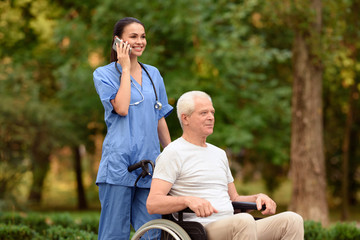 Nurse and old man sitting in a wheelchair in the park. Nurse talking on mobile phone