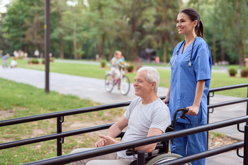 An old man in a wheelchair and a nurse strolling along a bridge in the park