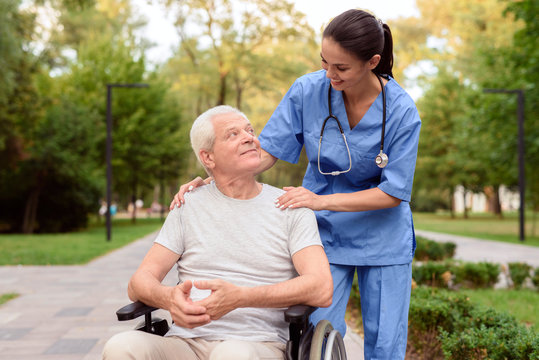 The nurse looks carefully at the old man, who is sitting in a wheelchair and smiling at him