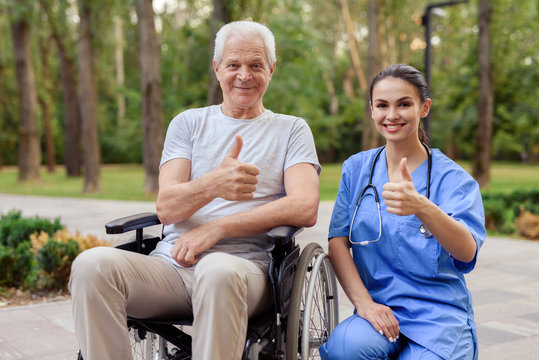 A nurse and an old man who sits in a wheelchair are showing a thumbs up