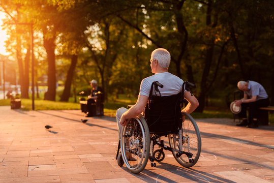 A lonely old man who sits in a wheelchair looks at a beautiful sunset in the park