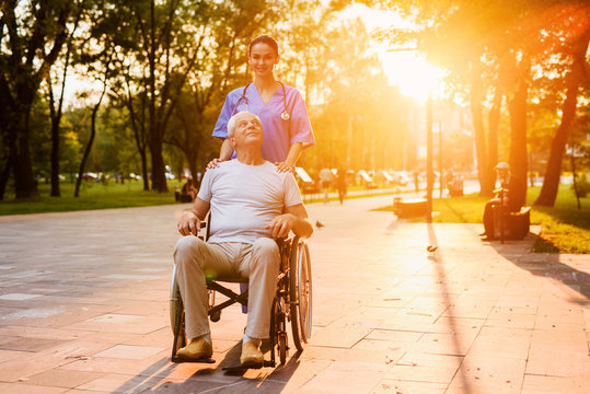 An old man who sits in a wheelchair and a nurse stand in the park at sunset