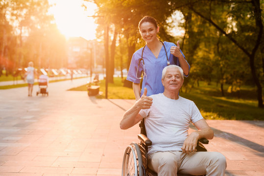 Nurse and old man who sits in a wheelchair showing a thumbs up in the park at sunset
