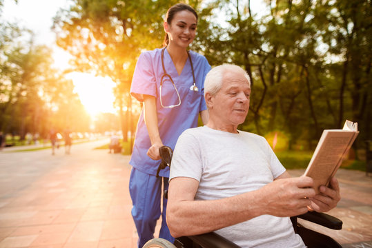 An old man who sits in a wheelchair and a nurse who is standing behind him reading a book in the park at sunset