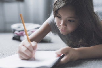 A girl lies on the gray floor next to the bed and draws something in her white notebook for drawing with a pencil