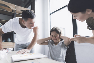 Obraz premium A man and a woman angrily scream at a small dark-haired girl who sits at a desk near the window, her hands over her ears