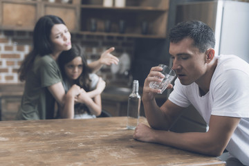 A man sits at a table and drinks, while a wife and daughter are sitting next to him and reproach him