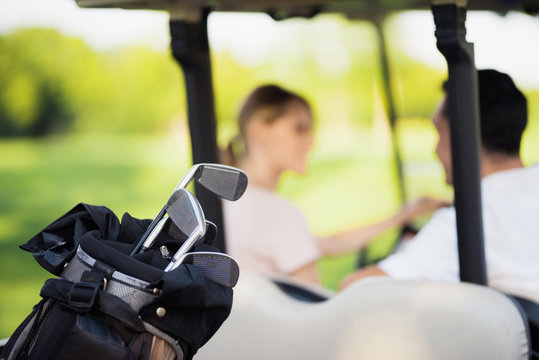 Close up photo. Golf clubs in the foreground, man with a woman in a golf cart on a background