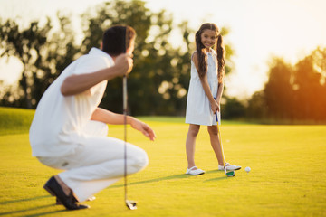 The father squats with a golf club in his hand and looks at his daughter, who looks at him and...
