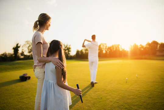 The man is looking into the distance, he just hit the ball with the club, the woman and the girl are looking at him