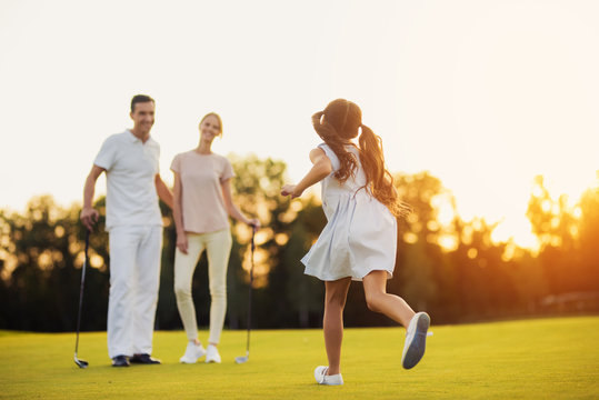 The girl runs across the golf course towards the man and woman who stand in front of her and golf clubs