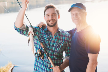 Obraz premium A man and an old man are posing with a fish that a man caught. Behind them, the sun is reflected in the river