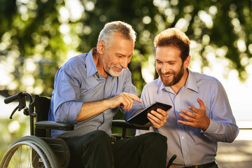 Old man in a wheelchair and a man looking at something in a tablet walking in the park. They are happy