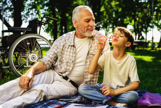 The old man and grandson are sitting on a picnic. The boy blows bubbles