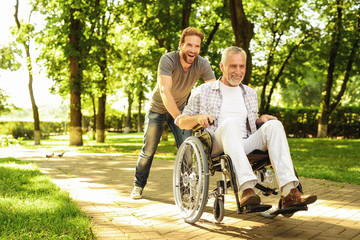 A pensioner on a wheelchair and his adult son are walking around the park. They are happy and have...