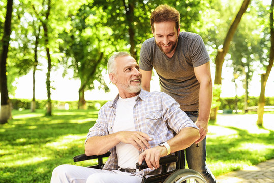 A man and his elderly father are walking in the park. A man carries his father in a wheelchair