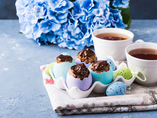 Easter sweet cup cakes treets in colorful pastel egg shells with chocolate and hydrangea flowers on blue background. Festive holiday home made food with cups of tea