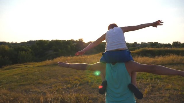 Little boy sitting on the shoulders of his father and playing airplane. Daddy carrying son and raised hands as plane. Family spending time together at nature. Slow motion Rear vack view