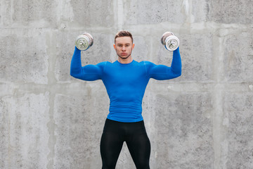 Muscular bodybuilder guy in a blue T-shirt doing exercises with dumbbells on the background of gray wall. Fitness and healthy lifestyle concept