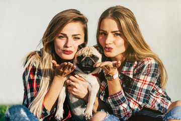 Two beautiful smiling sisters twins and dog pug sitting and sending an air kiss on green grass in...