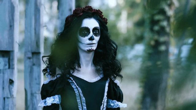 The Mexican Day of the Dead. The portrait of young woman with frightening make-up for Halloween standing in front of wooden old house and looking around. 4K