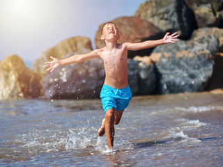 Cute Caucasian boy is running in the water along the sea shore. His arms are wide open.