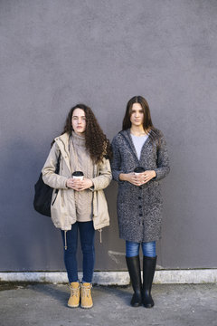 Two girlfriends standing against of grey wall outdoors